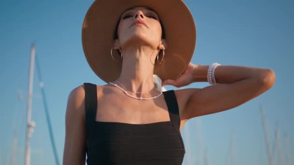 优雅的女人把草帽摆出美丽的蓝天 穿着时髦头饰站在夏天堤岸上的时髦年轻模特儿的画像 华丽的富家女看着阳光普照的自然 — 图库视频影像