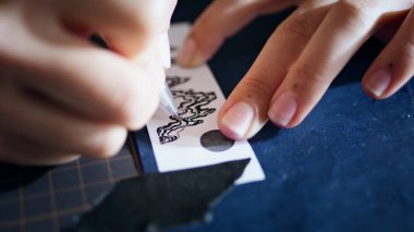 Dövmeci elleri yakın çekimde robot resmi aktarıyor. Ev stüdyosunda karbonik fotokopi kağıdıyla çalışan tanınmayan bir sanatçı. Kadın parmakları sadece yapıştırıcı bantla resim üzerine kalem çiziyor.