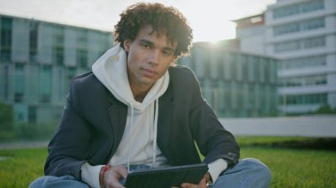 Çimen portresinde tablet tutan genç adam. Kıvırcık çocuk üniversite ortamında rahatlayan kameraya yakın çekim yapıyor. Yakışıklı, dokunaklı alet ekranı. Güneş ışınları binasının arka planında oturuyor.