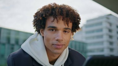 Şehir merkezindeki portrede genç bir adam var. Güler yüzlü Latin öğrenci, güneş ışığı mimarisi arka planında bir aletle oturuyor. Çağdaş kentsel bölgede poz veren pozitif genç yüzü