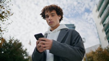 Genç adam açık havada telefon portresi kullanıyor. Kulaklıklar genç müzik dinleyen şehir arka planındaki kameralar yakınlaştırın. Güz sokağında tek başına poz veren akıllı telefonu elinde tutan yakışıklı adam.