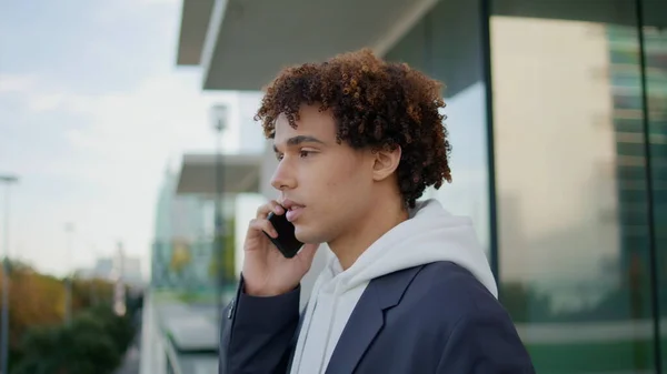 Young man having mobile call at glass balcony close up. Curly guy using phone speaking friend in modern architecture background. Handsome teenager holding cellphone talking calmly at terrace place