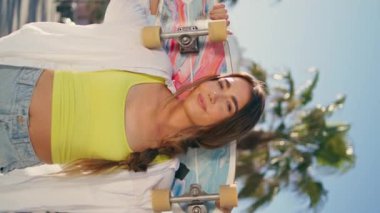 Gülümseyen kadın elinde kaykayla sokak resmine poz veriyor. Arka plandaki palmiye ağaçlarına bakan mutlu bayan. Yakın plan. Kendine güvenen kaykaycı kız, yaz tatilinin tadını çıkarıyor. Uzun tahtalı genç model. 