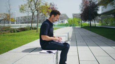 Akıllı iş adamı güneş ışınları caddesinde dizüstü bilgisayar kullanıyor. Serbest çalışan, internette sörf yapan bir bilgisayar. Esmer adam dijital cihazda çalışıyor beton bankta oturuyor. İnsan yaşam tarzı 