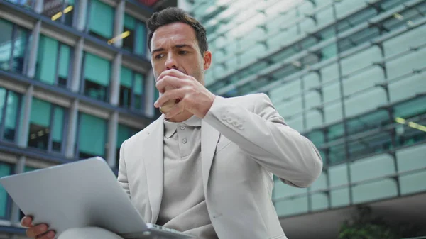 Leader boss drinking coffee sitting glass walls place closeup. Elegant hispanic businessman working laptop online sipping takeaway beverage zoom out. Confident man looking computer screen at open air