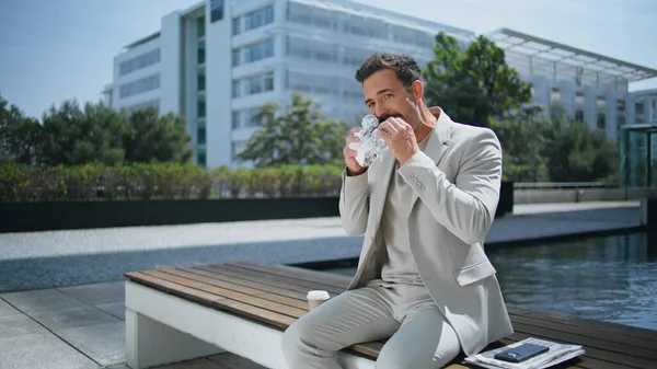 Relaxed businessman eating sandwich at wooden bench. Italian employee enjoying lunch break resting at greenery downtown. Formal suit groomed man tasting fast food at open air place. People lifestyle