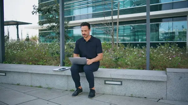 Hispanic manager talking by wireless earbuds working street. Casual smiling businessman sitting urban bench looking laptop computer screen. Elegant man consulting client solving company tasks remotely