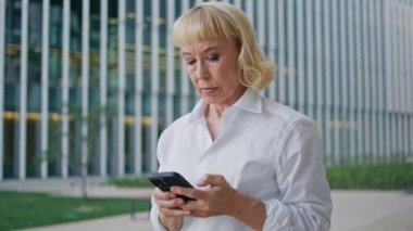 Emekli kadın CEO cep telefonuyla mesajlaşıyor. Şehir caddesinde yürüyor. Kapatın. Ciddi, yaşlı bir iş kadını akıllı telefon ekranı mesajı okuyor. Cep telefonuyla sosyal medyada gezinen lüks yaşlı kadın.