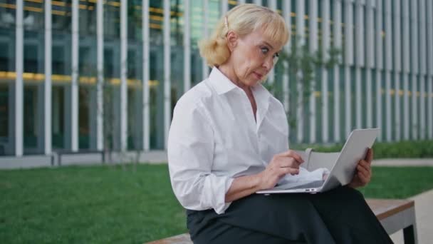 ストリートベンチでキーボードのノートパソコンをタイピングするプロのタイピング エレガントな金持ちの年金持ちは 屋外に座っているコンピュータ画面を見ていることに驚きました ワイヤレスガジェットでビジネスメールを読む成熟した女性 — ストック動画