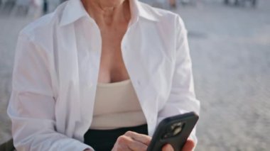 Güneşli şehirde rahat bir emeklinin cep telefonuna mesaj atması. Kırışık eller dışarıda mesaj yazarken akıllı telefon ekranına dokunuyor. Gülümseyen emekli kadın sosyal medyada geziniyor, sokak ortasında oturuyor..