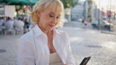 Gülümseyen yaşlı bir kadın cep telefonuna cep telefonuyla mesajlaşıyor. Güzel buruşuk kadın yüzü restoran masasındaki akıllı telefon ekranına bakıyor. Rahatlamış kadın emekli dışarıda mesaj okuyor.