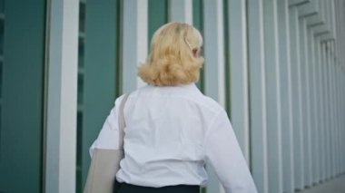 Modern şehir bölgesinde yürüyen bir emeklinin arkası. Beyaz tişörtlü, tanınmayan yaşlı kadın çağdaş ofis binasının yakınına gidiyor. Emekli sarışın, iş kadını, şehirde geziyor..