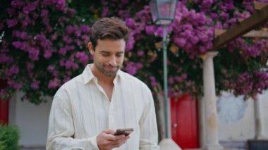Rahatlamış centilmen çiçek sokağında geziniyor. Yakın plan. Memnuniyet duyulan mutlu adam elinde akıllı telefon tutup şehir balkonunda mesaj okuyor. Latin sakallı maço tatilde cep telefonuyla internette geziniyor.