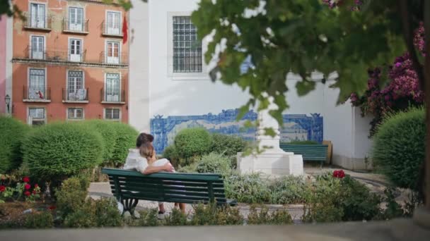 優しく座っている公園のベンチのバックビューを抱擁するロマンチックなペア ブルネット男 市のデートでガールフレンドと絆を結ぶ 緑の広場の休暇で一緒に時間を楽しむカップルを魅了しました ラブコンセプト — ストック動画