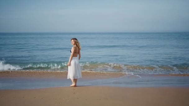 落ち着いた泡立った波を一人で歩く海岸をリラックスした女性 濡れた砂の上を散策するリラックスしたカーリーモデルが美しい海岸を眺めます キャリーフリーロマンチックな女性観光客は晴れた夏の週末を楽しむ — ストック動画