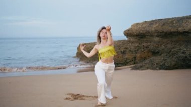 Okyanus dalgalarının önünde dans eden kıvırcık kız. Profesyonel bayan dansçı, kum plajında modern stilde hareket eden esnek bir vücut. Çağdaş sanatçı kadın, doğada duygusal bir performans sergiliyor..