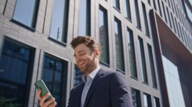 Pozitif yönetici akıllı telefondan mesaj okuyor. Güneşli bir sabahta işe gidiyormuş. Yakın çekim. Cep telefonu ekranına gülen yakışıklı bir adam. Neşeli CEO şehir merkezinde yürüyor. Mesajlaşma telefonu..