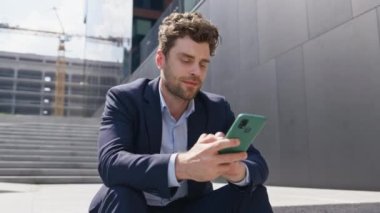 Neşeli, serbest çalışan cep telefonu görünümlü, gülümseyen şehir merdivenlerinde oturan bir iş yeri. Yakışıklı iş adamı dışarıdaki akıllı telefondan haber aktarıyor. Rahatlamış adam, tatilin tadını çıkar..