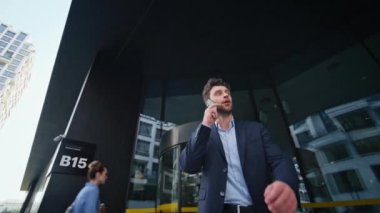 Memnun iş adamı cep telefonundan modern ofis binasından ayrılıyor. Mutlu menajer telefonla harika haberler aldığı için heyecanlanıyor. Gülümseyen duygusal adam akıllı telefonla konuşuyor..