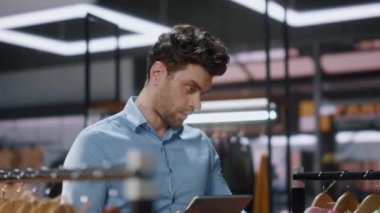 Modern giyim mağazasında elinde tablet tutan sıralı bir adam. Mutlu butik işçisi müşterilere gülümseyerek envanter çıkarıyor. Alışveriş merkezinde çalışan kendine güvenen mağaza asistanı dijital aygıtı kontrol ediyor
