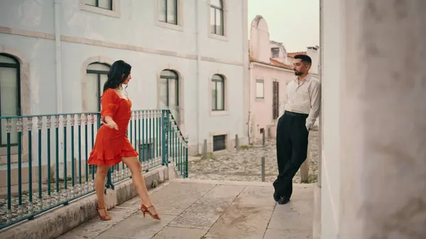 Beautiful bright woman dancer flirting with elegant partner starting dance near building railings. Attractive girl performer in red dress approaching to man leaned at wall. Passionate couple dancing.
