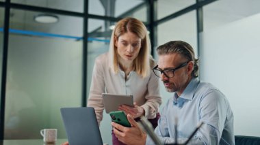 Kabine kapanışında iş konuşmaya odaklanmış iş arkadaşları. Bayan asistan ofisteki ciddi iş adamına tablet gösteriyor. Akıllı telefondan dizüstü bilgisayarı işaret eden meşgul bir adam. Motivasyonlu ekip beyin fırtınası 