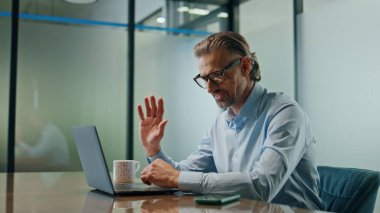 Gülümseyen menajer kapalı mekandaki video çağrısını selamlıyor. Dost canlısı lider iş yerindeki bilgisayar görüntülü sohbete el sallıyor. Modern iç mekandaki pozitif patron merhaba diyor. 