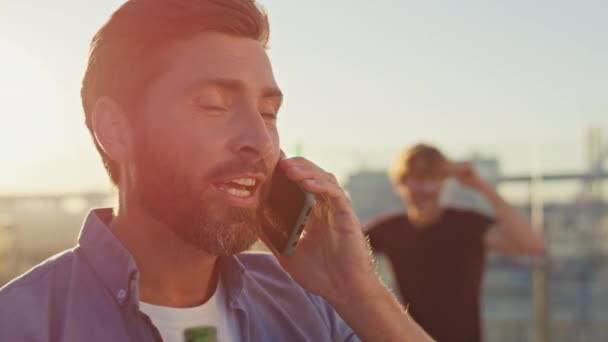 夏の屋上クローズアップでスマートフォンを呼ぶパーティーマン 笑顔のひげをした男は 夜の日光にビール瓶を持っている携帯電話を話します ハッピーリラックスしたヒップスターは 友達のハングアウトで電話会話を持っています — ストック動画