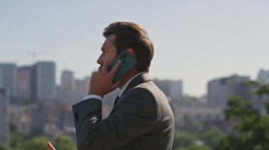 Sakallı yönetici caddede cep telefonuyla konuşuyor. Kendine güvenen işadamı, şık takım elbise içinde güneşli şehir panoramasında duran ortakları arıyor. Varlıklı adam iş konuşuyor. Kamera hareket ediyor