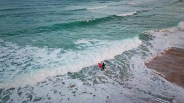 Okyanusa giren bilinmeyen sörfçü fırtınalı dalgalarda havada sörf tahtası tutuyor. Kıyı şeridine doğru ilerleyen derin turkuaz deniz köpüğü süper yavaş çekimde. Yazın kumsalda süzülen mavi su köpüklü deniz kıyısı