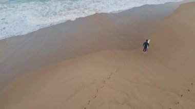 Sörfçü adam kumsalda yürürken ayak izleri bırakıyor. İHA sörf tahtası taşıyan sörfçü ıslak kumu köpüklü okyanus suyuyla yavaş çekimde basıyor. Harika bir deniz manzarası. Aktif hobi eğlence konsepti 