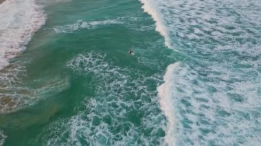 Yazın kumsalda yuvarlanan derin mavi okyanus köpüğü. Sörf tahtasında uzanmış fırtınalı dalgaları bekleyen tanınmamış bir sörfçü. Kıyı şeridine doğru ilerleyen turkuaz deniz köpüğü.