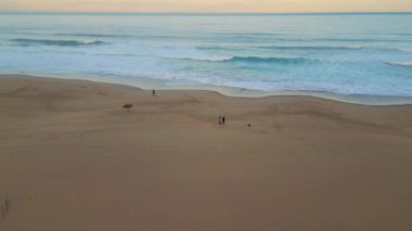 Gün batımını kaplayan okyanus sörfü drone görüntüsü. Deniz yüzeyine yansıyan turkuaz dalgalar plajdaki altın renkli pastel güneş ışığını kaplıyor. İnsanlar deniz kıyısında yürüyen siluetleri, sahil şeridi gündoğumu havasını yıkıyor.