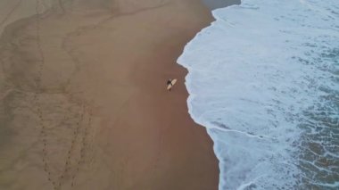 Drone, kumsalda yürüyen ve dalgaları bekleyen bilinmeyen sörfçüyü vurdu. Deniz manzaralı harika sörfçü sörf tahtası taşıyor ıslak kumu köpüklü okyanus suyuyla yavaş çekimde basıyor. Aktif hobi eğlence konsepti 