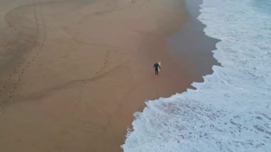 Yalnız sörfçü sahil drone manzarasında yürüyor. Kasvetli bir günde kıyıları kaplayan köpüklü okyanus dalgaları. Yuvarlanan deniz suyu, yıkanan kumlu yüzey. Tanımlanamayan sporcu hafta sonları panoda bekliyor.