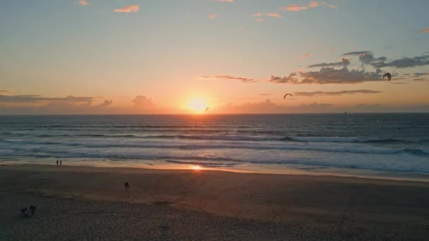 海のサーフィン 日の出を洗う 海岸の空中ビュー 空中を飛ぶビーチパワーキットをカバーするファミリー波 海面を反射する暖かい太陽光線 人々はキットサーフィンスポーツを見ている海岸を歩いているシルエット — ストック動画