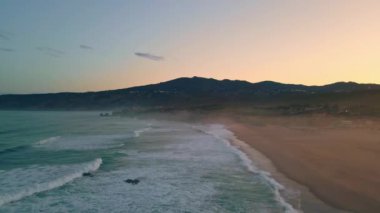 Gündoğumu deniz sörfü doğa drone görüntüsü. Bej rengi kumları yıkayan ışık dalgalarıyla birlikte sakin bir sabah okyanusu. Pastel gökyüzü turkuvaz su muhteşem manzara. Arka planda karanlık dağlar