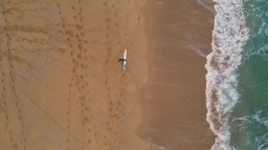 Sörf tahtası manzaralı, kumsalda yürüyen sörfçü. Güzel okyanus suyunun köpürmesi çok yavaş bir şekilde sakin bir sahile vuruyor. Köpüklü deniz dalgalarında sörf yaptıktan sonra tanınmayan biri rahatlıyor..