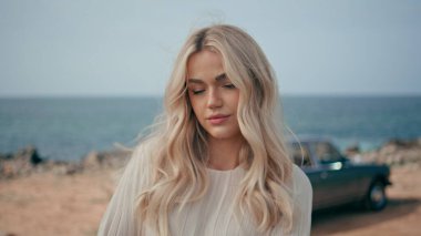 Yakından bakınca güzel, genç bir model güzel bir yaz deniz kıyısında, retro arabanın önünde duruyor. Okyanus kıyısında poz veren çekici sarışın kadın. Kameraya özgüvenle bakıyor. Güzel kız gezinin tadını çıkarıyor..