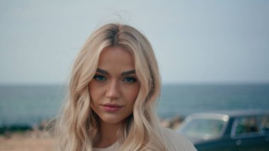 Lüks sarışın kızın portresi. Kameraya özgüveni tam görünüyor. Arka planında retro arabasıyla güzel bir deniz kıyısında duran muhteşem genç kadın. Okyanus kıyısında otomatiğe yakın poz veren havalı bir model.