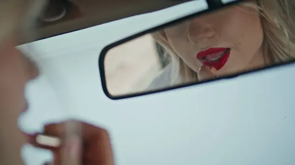 Car mirror reflecting beautiful woman applying red lipstick on sensual lips close up. Gorgeous blonde making makeup sitting inside automobile. Young attractive girl looking on face beauty reflection.