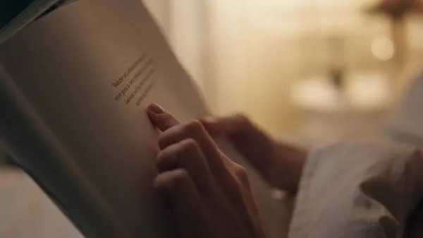 Closeup hands holding book indoors. Smiling peaceful woman reading favourite novel on weekend holiday evening. Calm girl student studying learning information in bed home. Finger pointing page lines