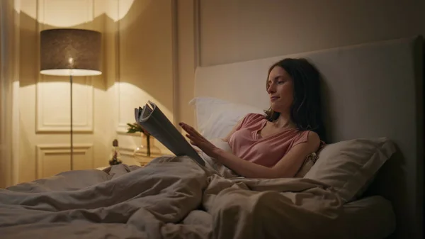 Evening woman reading novel in cozy bed. Smiling relaxed reader resting home enjoying book before sleep. Focused student turning page studying late in lamp light. Insomnia leisure lifestyle concept.