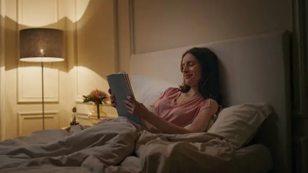Peaceful woman reading book in evening home. Smiling reader enjoying novel rest in cozy bed on weekend night. Calm relaxed female chill studying late in lamp light. Favourite literature hobby concept.