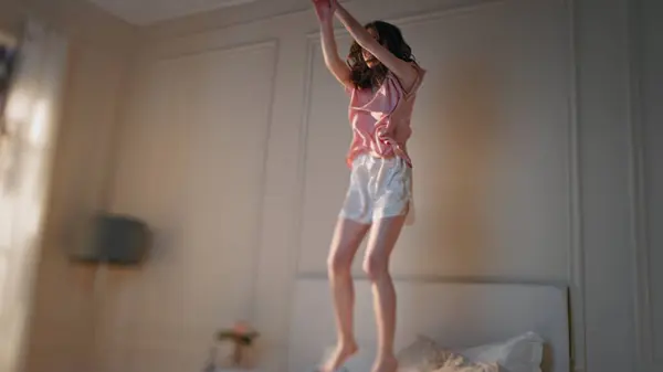 Excited woman jumping bed in morning. Happy beautiful girl dance home in pajamas moving to energetic music playlist. Joyful slim model relaxing enjoying time alone. Smiling female celebrating freedom