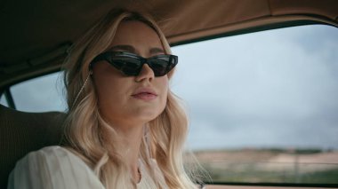 Rahat bir eski model arabayla seyahat eden kadın sürücünün portresi. Bulutlu havada araba kullanan çekici genç bir gezgin kız. Güneş gözlüklü şık sarışın. Dümende kamera var.