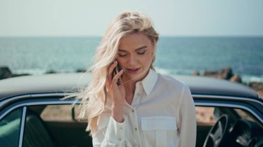 Sarışının portresi akıllı telefon konuşuyor. Retro arabanın yanında güzel bir deniz kıyısında duruyor. Çekici, rahat, cep telefonlu bir kadın okyanus kıyısında yaz tatilinin tadını çıkarıyor. Çok güzel bir kız, çok yakından konuşuyor.