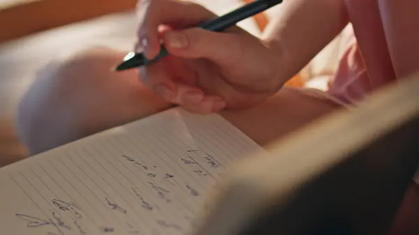 Hand making handwriting notes closeup. Girl holding pen planning day in morning. Unrecognized woman arm closing book in golden sunlight. Dreamy romantic female writing diary enjoy weekend in pajamas.