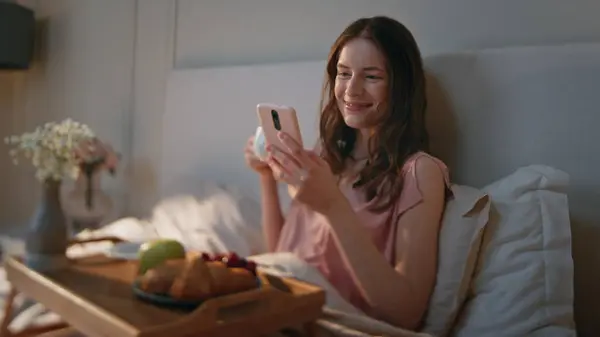 Joyful woman scrolling social media in bed closeup. Happy attractive girl rest enjoying breakfast coffee at home. Carefree smiling female using mobile phone watch funny content. Lifestyle fun concept