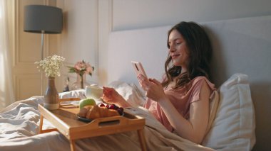 Otel odasında akıllı telefon kullanan barışçıl bir kadın. Sakin kadın elinde kahve fincanıyla yatakta kahvaltının tadını çıkarıyor. Çekici gülümseyen kız hafta sonu internette telefon alışverişi yapıyor. Uyumlu zihinsel denge kavramı
